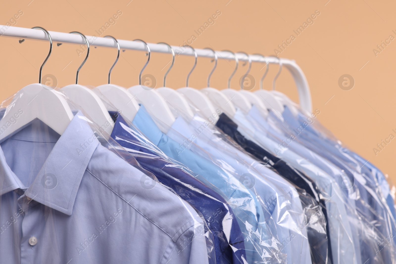 Photo of Dry-cleaning service. Many different clothes in plastic bags hanging on rack against beige background, closeup