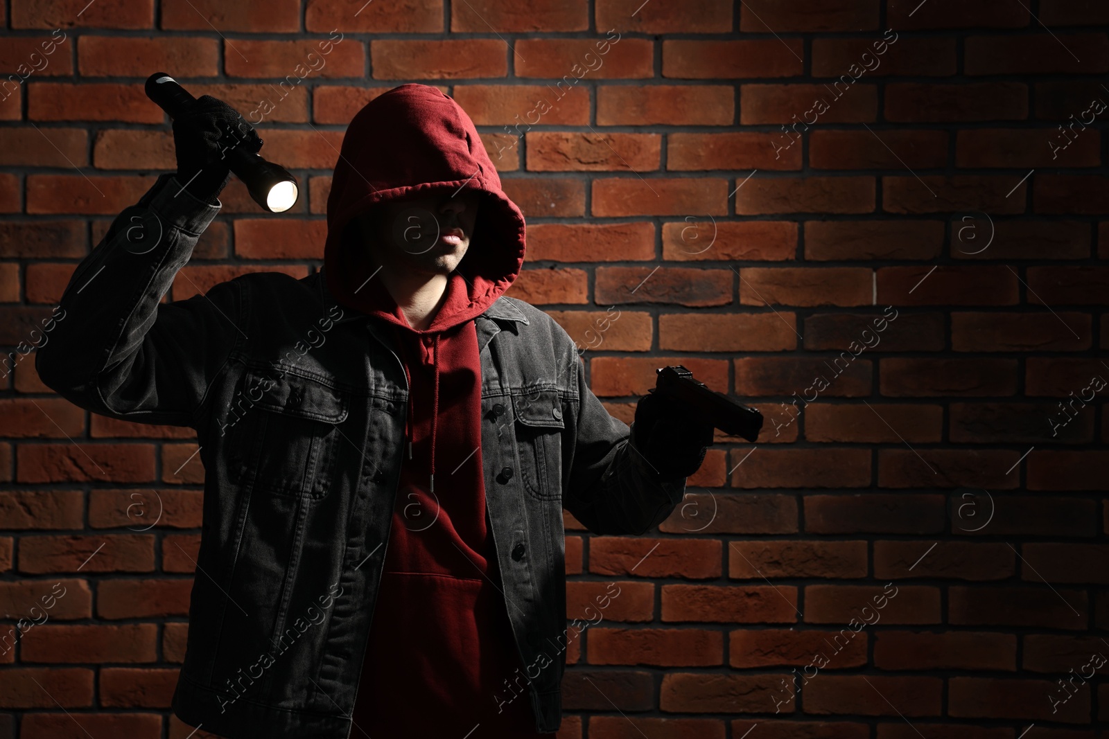 Photo of Thief in hoodie with gun and flashlight against red brick wall