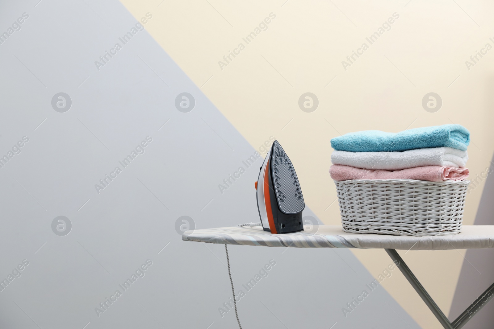 Photo of New modern iron and basket with laundry on board against color background, space for text