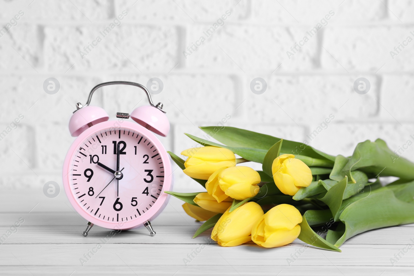 Photo of Pink alarm clock and beautiful tulips on white wooden table against brick wall. Spring time