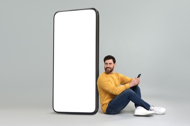 Image of Man with mobile phone sitting near huge device with empty screen on grey background. Mockup for design