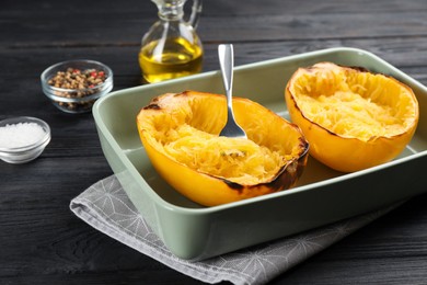 Photo of Halves of cooked spaghetti squash and fork in baking  dish on black wooden table, closeup
