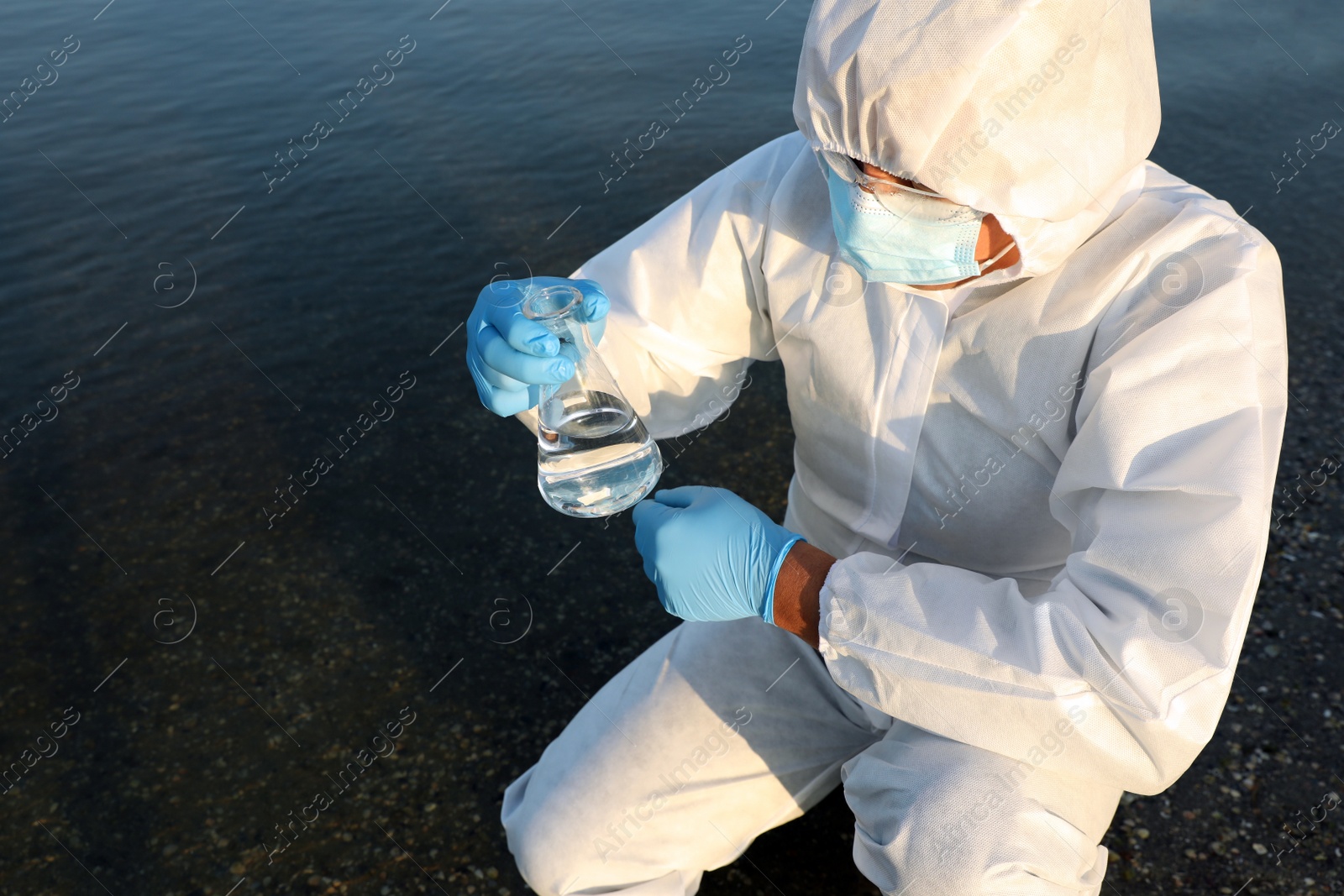 Photo of Scientist in chemical protective suit with conical flask taking sample from river for analysis