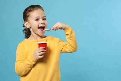 Girl eating tasty yogurt on light blue background, space for text