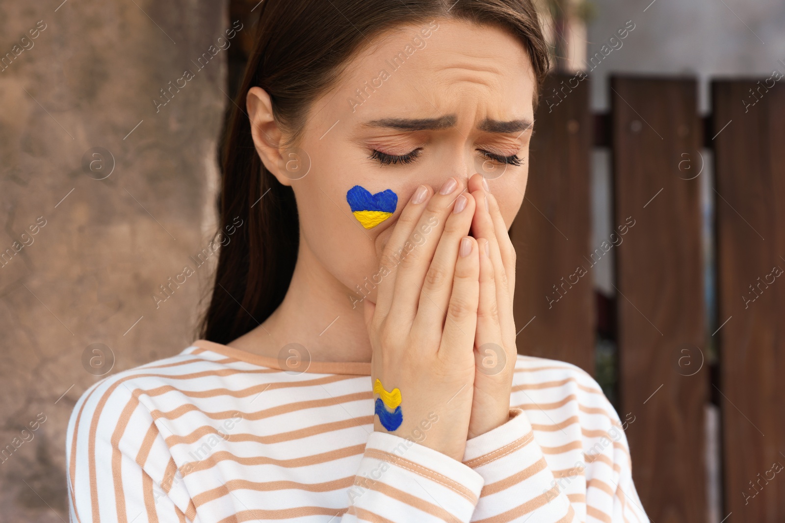 Photo of Sad young woman with drawings of Ukrainian flag on face outdoors, closeup
