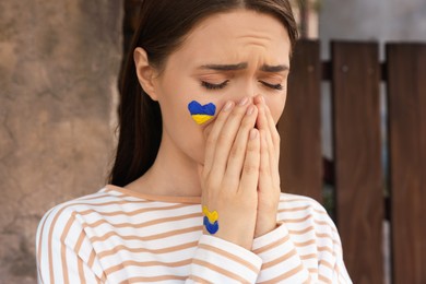 Photo of Sad young woman with drawings of Ukrainian flag on face outdoors, closeup