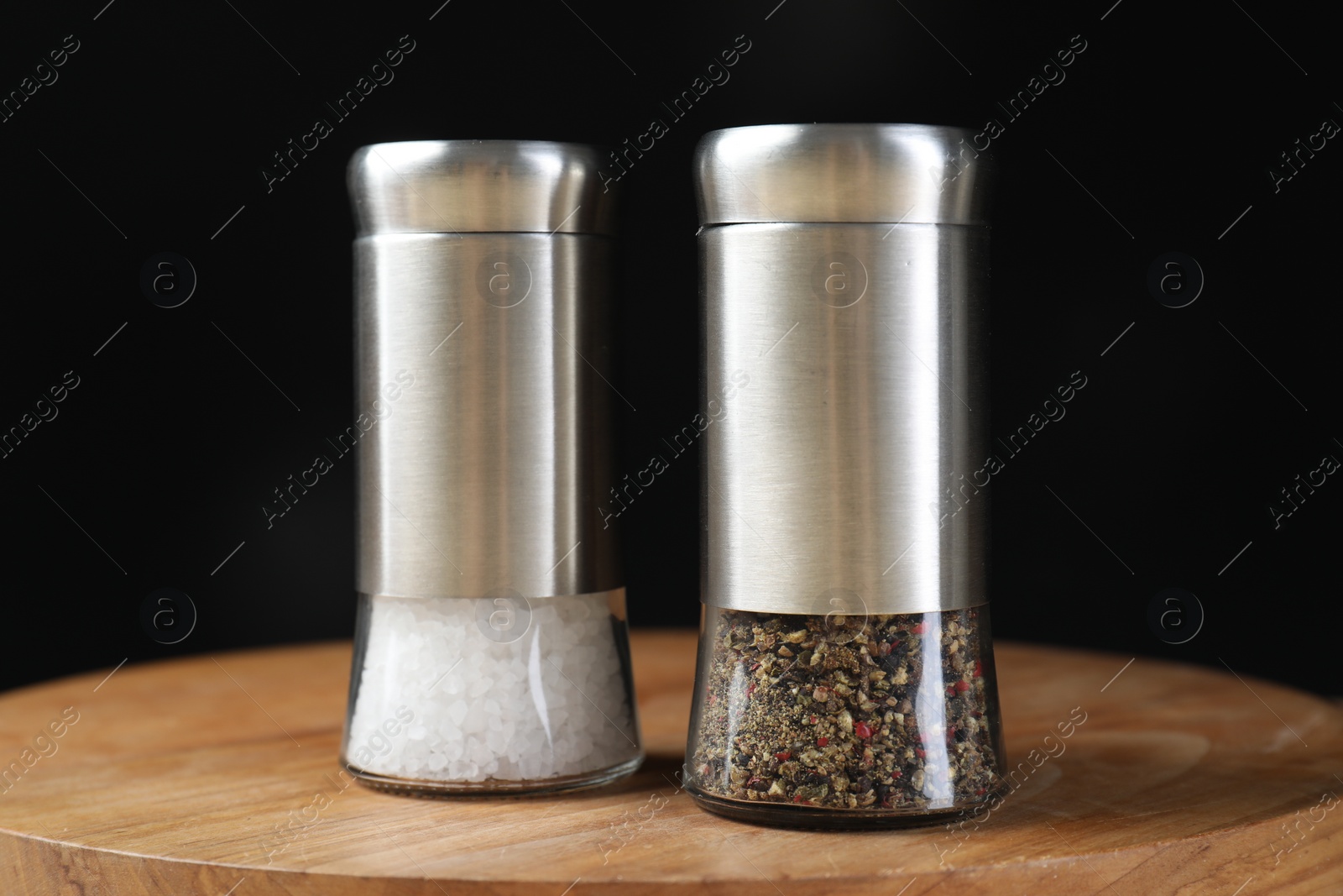 Photo of Salt and pepper shakers on wooden board against black background, closeup