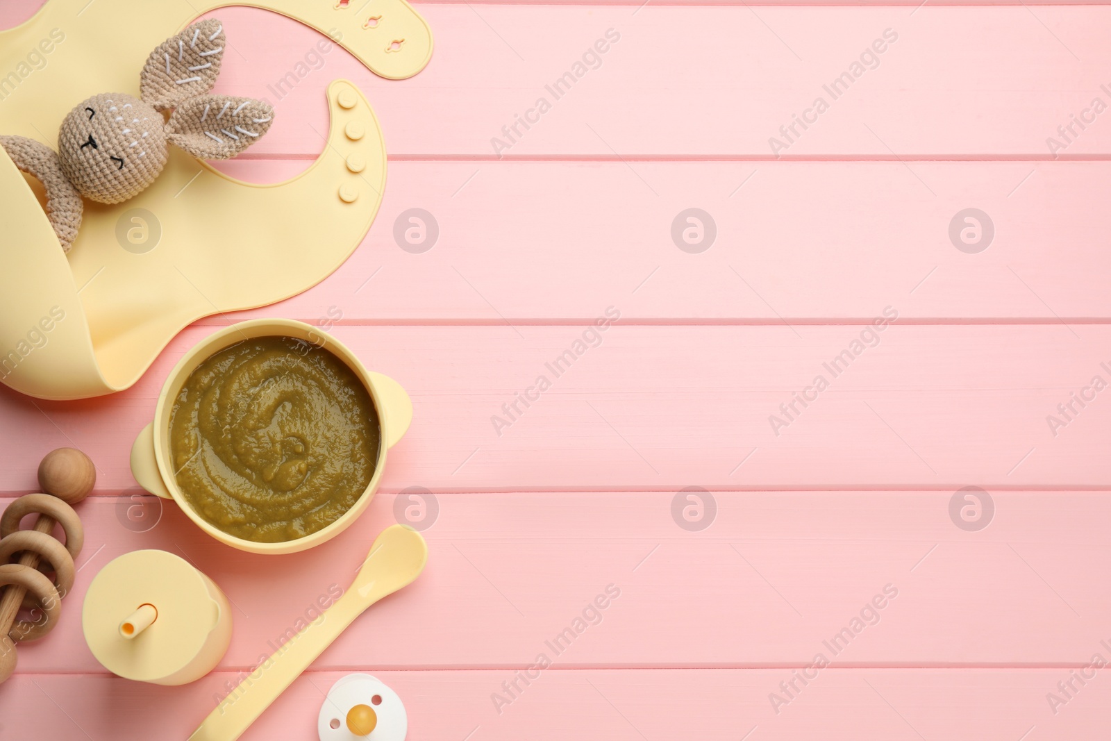 Photo of Silicone baby bib, toys and plastic dishware with healthy food on pink wooden table, flat lay. Space for text