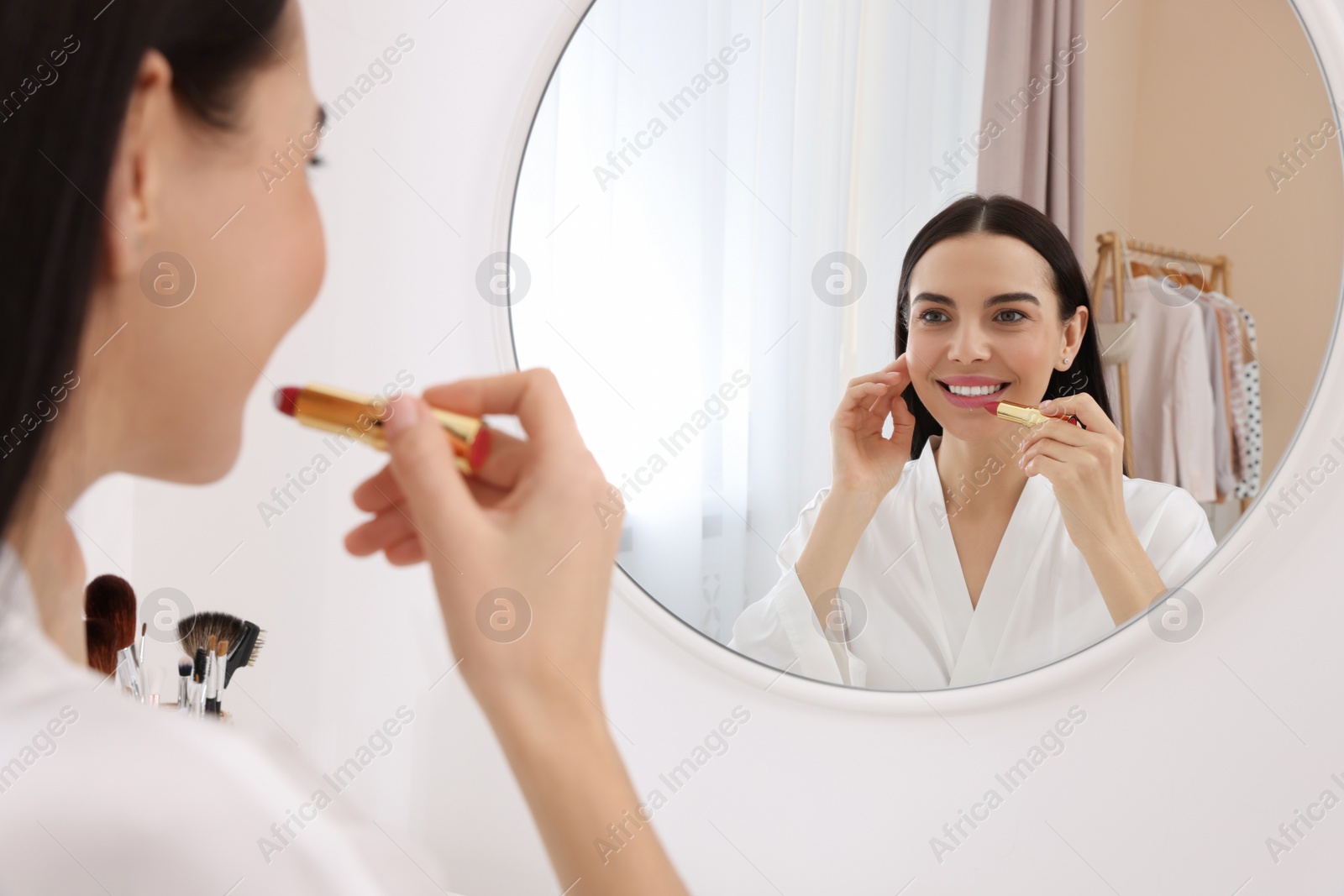 Photo of Beautiful young woman applying lipstick near mirror indoors