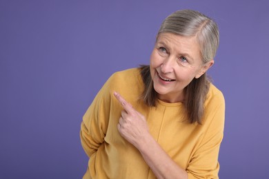 Surprised senior woman pointing at something on violet background, space for text
