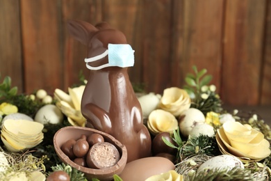 Photo of Chocolate bunny with protective mask, eggs and wreath against wooden background, closeup. Easter holiday during COVID-19 quarantine