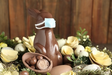 Chocolate bunny with protective mask, eggs and wreath against wooden background, closeup. Easter holiday during COVID-19 quarantine