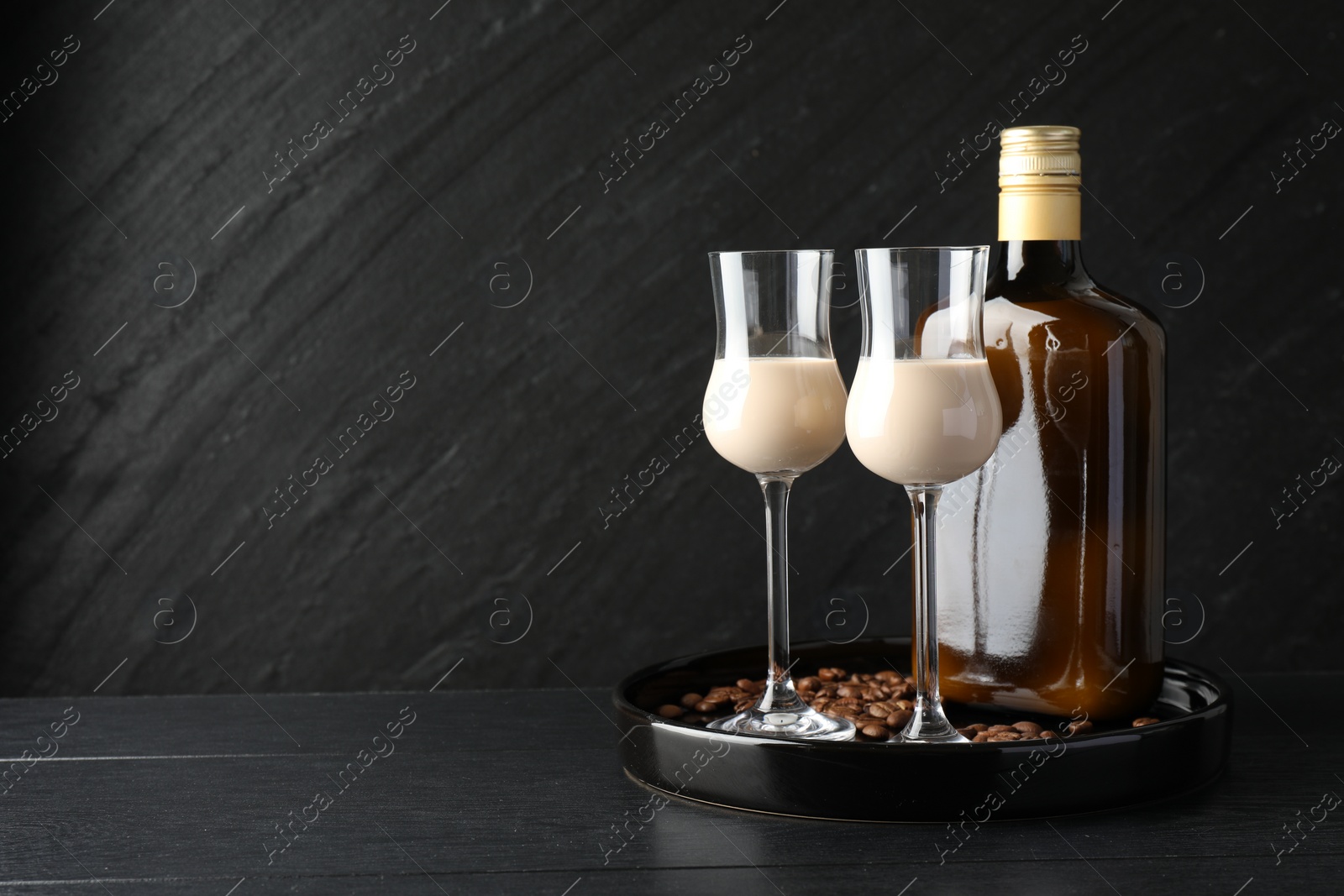 Photo of Coffee cream liqueur in glasses, bottle and beans on black wooden table, space for text
