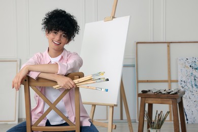 Photo of Young woman holding brushes near easel with canvas in studio