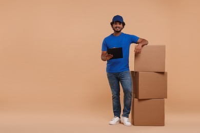 Photo of Happy courier with clipboard and stack of parcels on light brown background, space for text