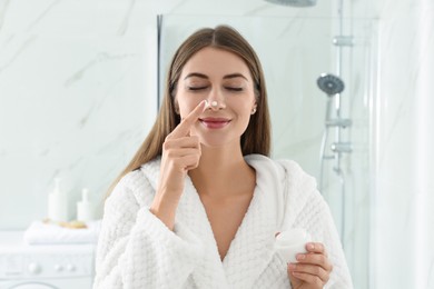 Photo of Young woman applying face cream onto her nose in bathroom