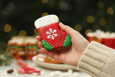 Woman with decorated cookie at table against blurred Christmas lights, closeup