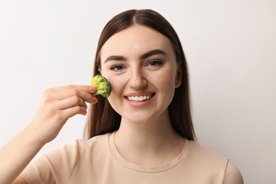 Photo of Smiling woman making fake freckles with broccoli and cosmetic product on light background