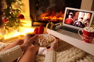 MYKOLAIV, UKRAINE - DECEMBER 23, 2020: Woman with cookies watching Harry Potter and Philosopher's Stone movie on laptop near fireplace at home, closeup. Cozy winter holidays atmosphere