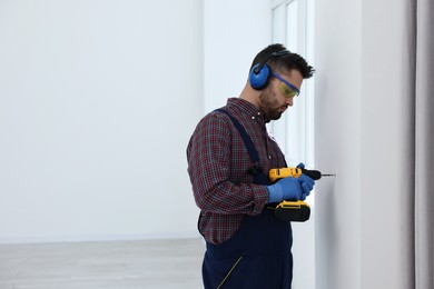 Photo of Young worker in uniform using electric drill indoors, space for text