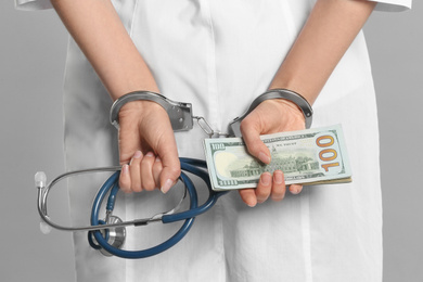 Photo of Doctor in handcuffs with bribe and stethoscope on grey background, closeup. Corrupted medicine