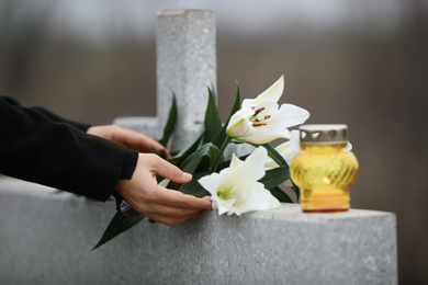 Woman holding white lilies near light grey granite tombstone with candle outdoors, closeup. Funeral ceremony