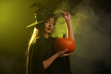 Photo of Young woman wearing witch costume with pumpkin in smoke cloud on dark background. Halloween party