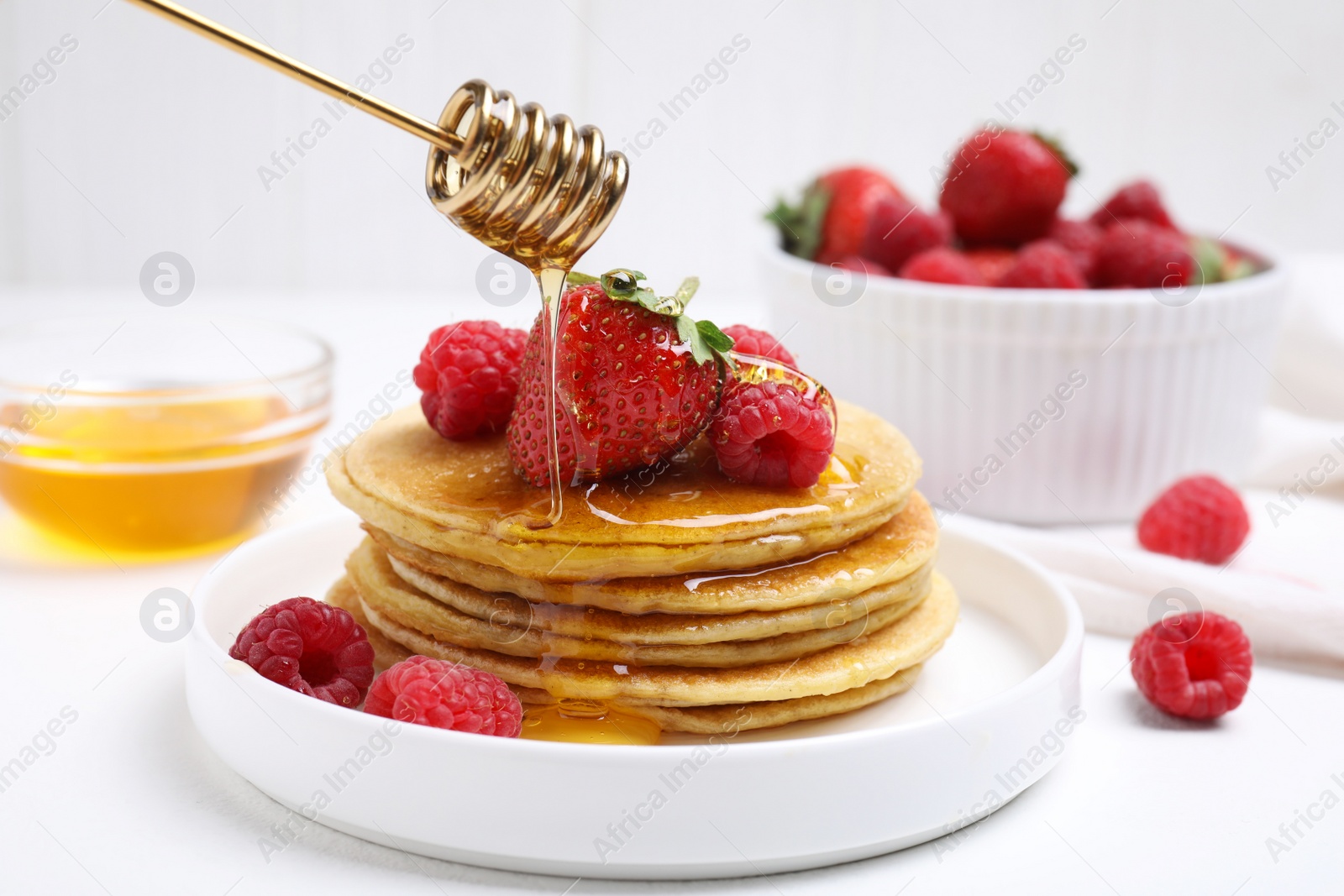 Photo of Pouring honey onto tasty pancakes with fresh berries on white table