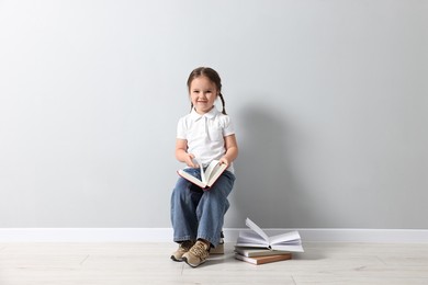 Cute little girl sitting on stack of books near light grey wall