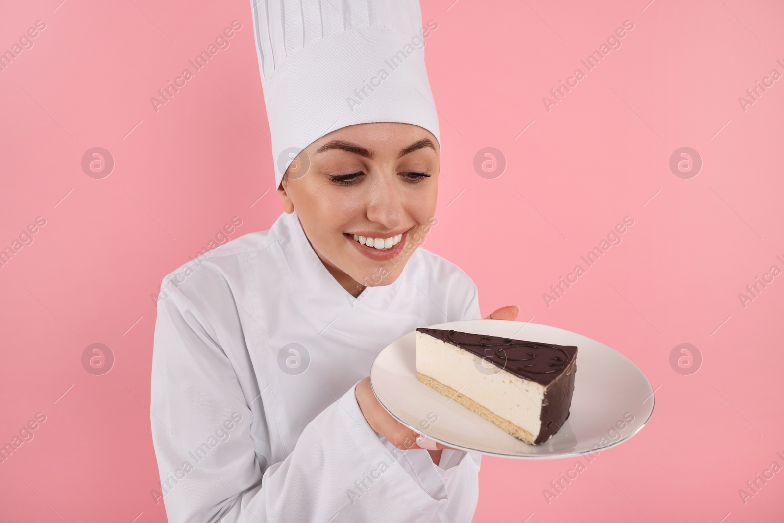 Photo of Happy professional confectioner in uniform holding delicious cheesecake on pink background