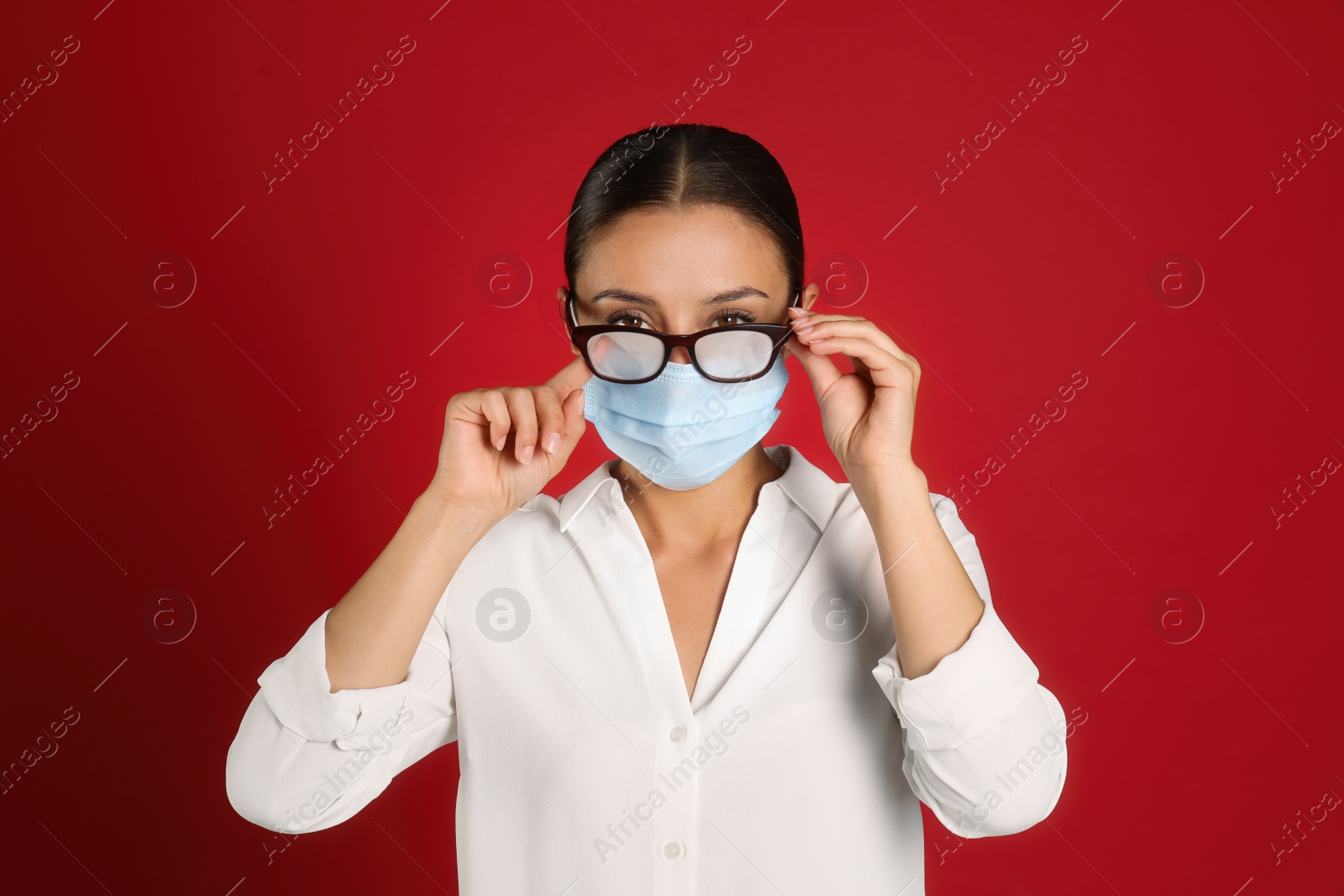 Photo of Woman wiping foggy glasses caused by wearing medical mask on red background