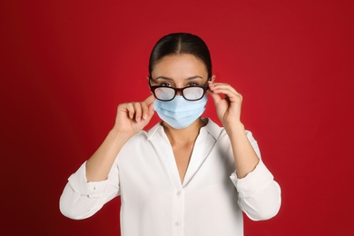 Woman wiping foggy glasses caused by wearing medical mask on red background