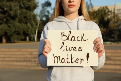 Photo of Young woman holding sign with phrase Black Lives Matter outdoors, closeup. Racism concept