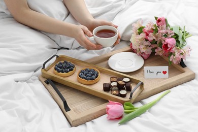 Photo of Tasty breakfast served in bed. Woman with tea, desserts, flowers and I Love You card at home, closeup