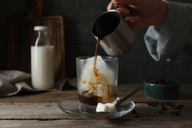 Woman pouring coffee into glass with ice cubes at wooden table, closeup