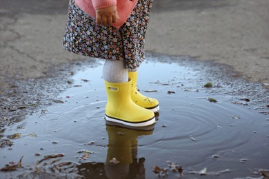 Photo of Little girl wearing rubber boots standing in puddle outdoors, closeup. Autumn walk