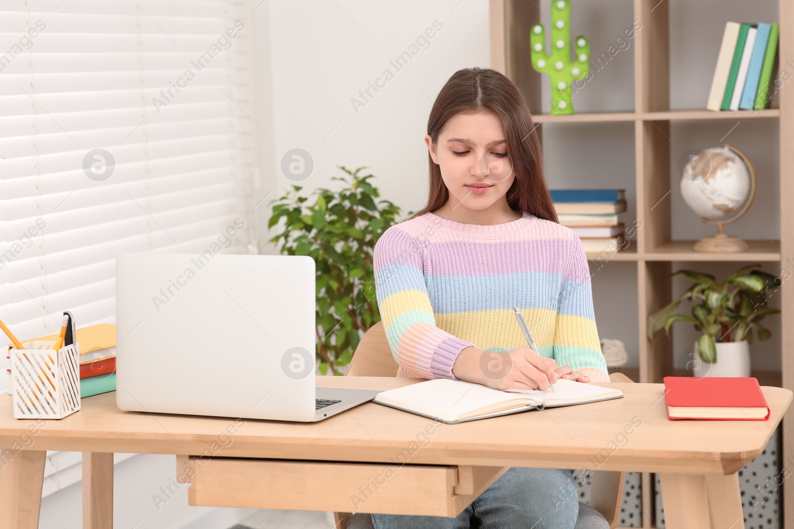 Photo of Cute girl writing in notepad near laptop at desk in room. Home workplace