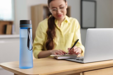 Woman working at wooden desk indoors, focus on bottle. Space for text