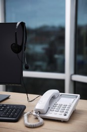 Photo of Stationary phone near modern computer with headset on wooden desk indoors. Hotline service
