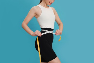 Photo of Happy woman in sportswear measuring waist with tape on light blue background, closeup