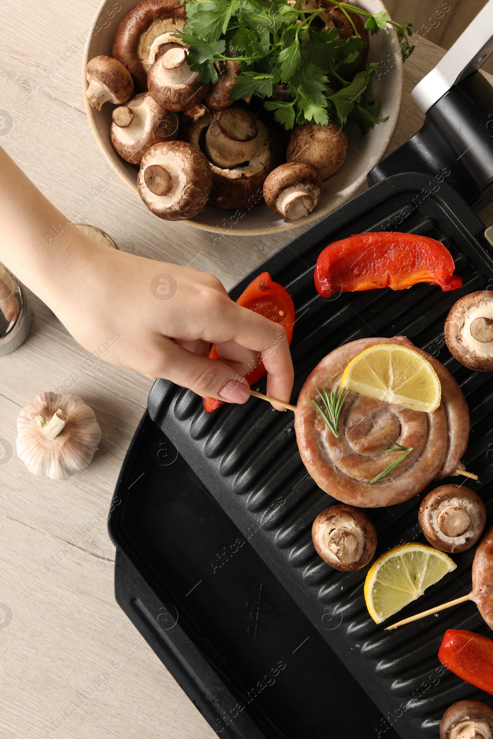 Photo of Woman cooking homemade sausages with mushrooms and bell pepper on electric grill at wooden table, top view