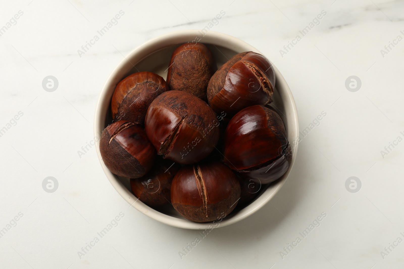 Photo of Roasted edible sweet chestnuts in bowl on white marble table, top view
