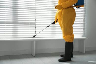 Photo of Male worker in protective suit spraying insecticide on window sill indoors, closeup. Pest control