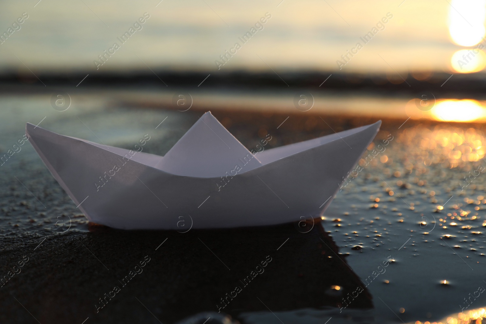 Photo of White paper boat on wet sand near sea at sunset