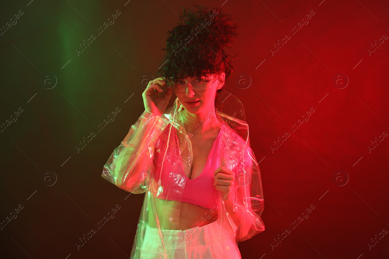 Photo of Beautiful young woman in transparent coat and sunglasses posing on color background in neon lights