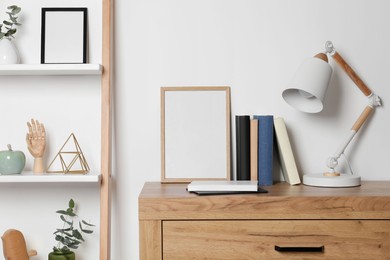 Photo of Stylish modern desk lamp, books and frame on wooden chest of drawers near white wall indoors