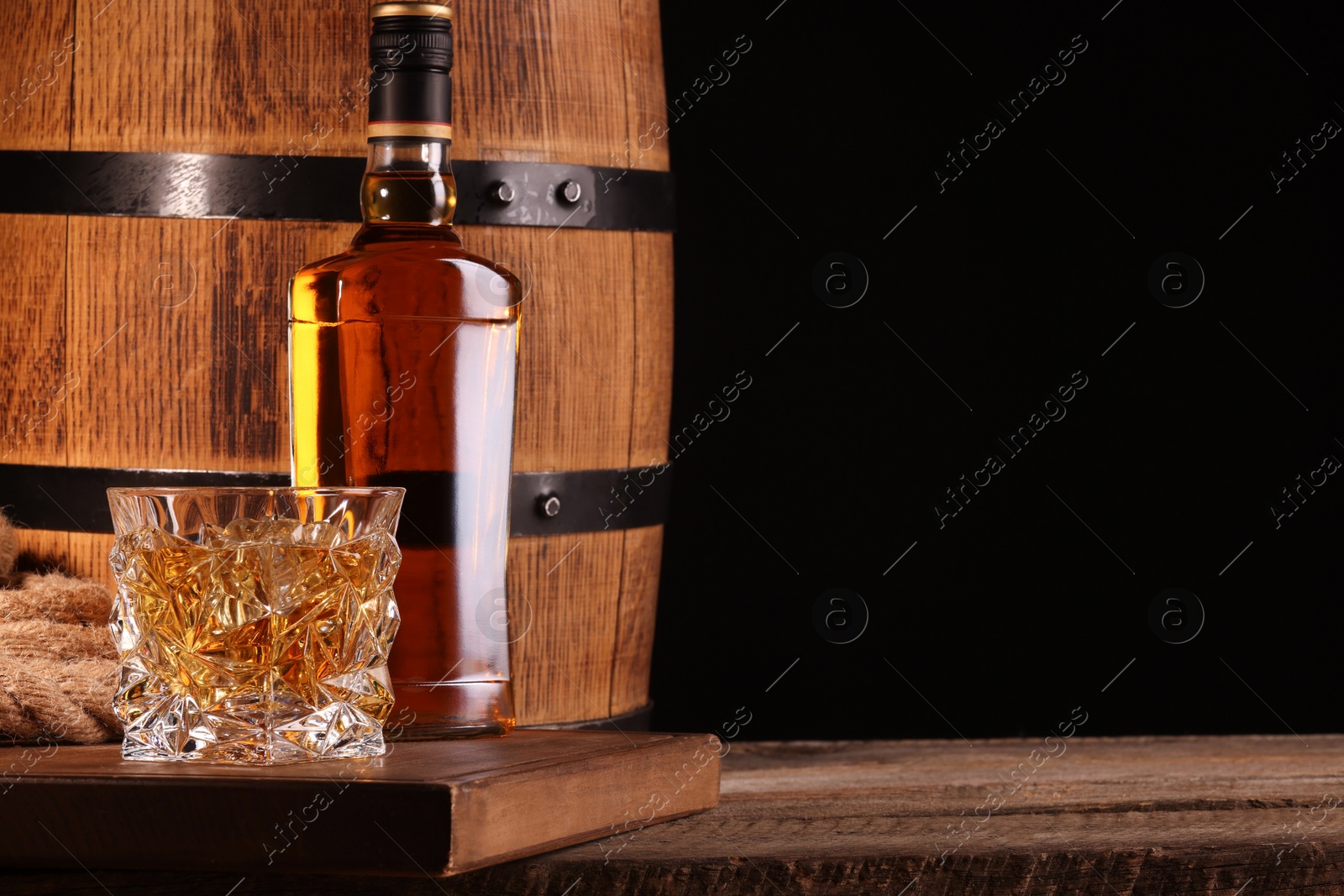 Photo of Whiskey with ice cubes in glass, bottle and barrel on wooden table against black background, space for text