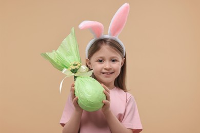 Photo of Easter celebration. Cute girl with bunny ears holding wrapped gift on beige background