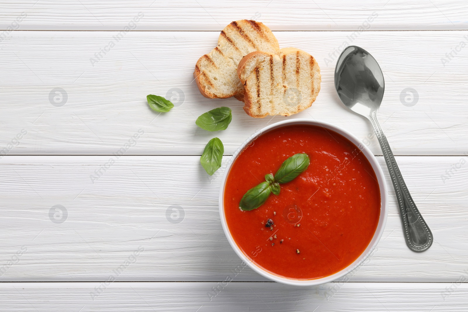 Photo of Delicious tomato cream soup in bowl served with pieces of grilled bread on white wooden table, flat lay. Space for text