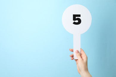 Woman holding auction paddle with number 5 on light blue background, closeup. Space for text